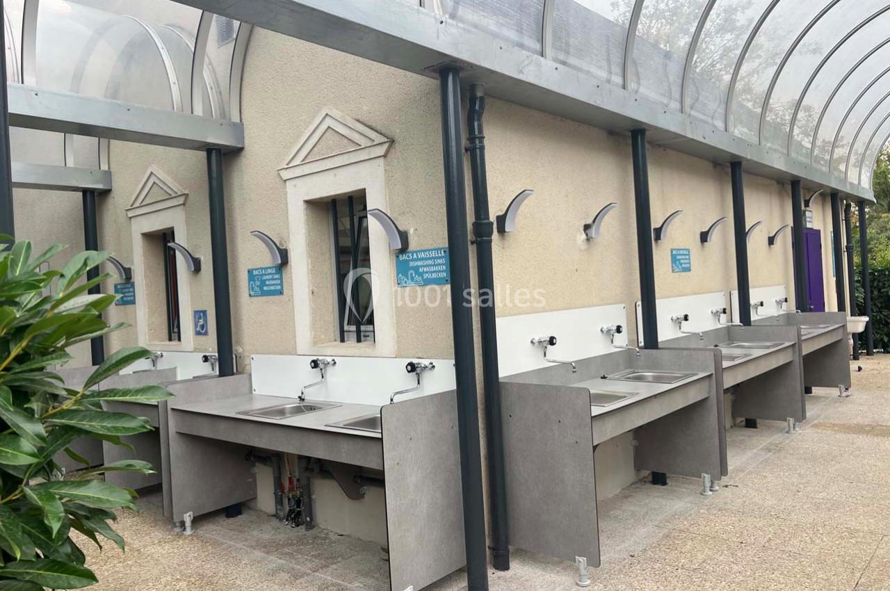 Batterie d'éviers extérieurs en inox sous un auvent transparent, adossés à un mur beige avec panneaux d'instructions.