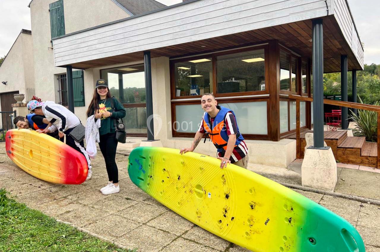 Deux personnes tiennent des planches de paddle devant un bâtiment, une troisième les observe en arrière-plan.