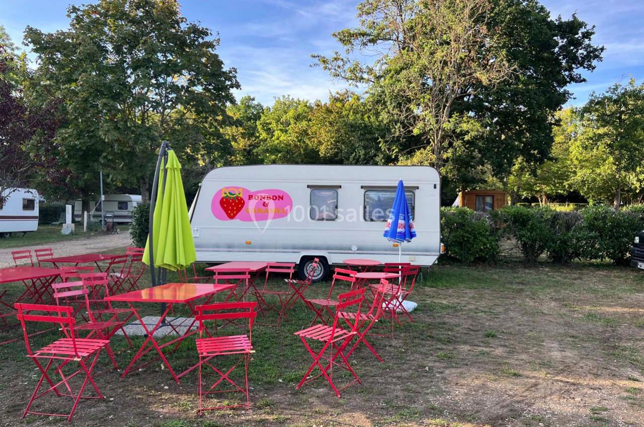 Caravane blanche avec un logo de fraise, entourée de tables et chaises rouges dans un espace extérieur arboré.