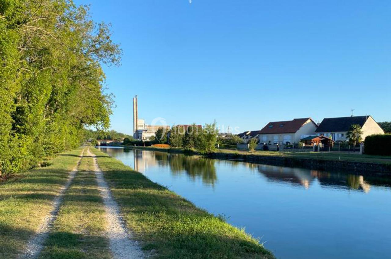 Chemin de terre longeant un canal bordé d'arbres à gauche et de maisons à droite, sous un ciel dégagé.