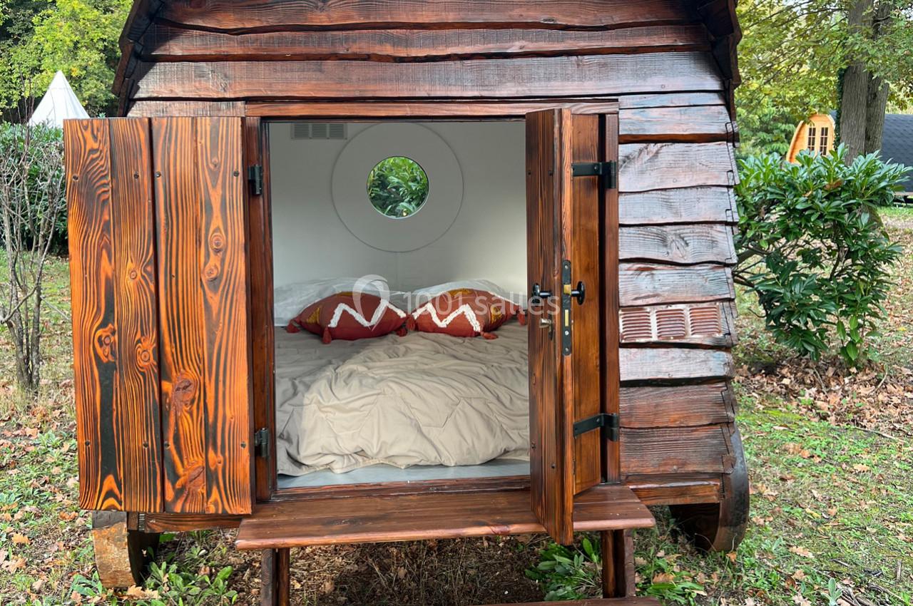 Cabane en bois avec portes ouvertes, révélant un lit double et un intérieur blanc, située dans un environnement naturel.
