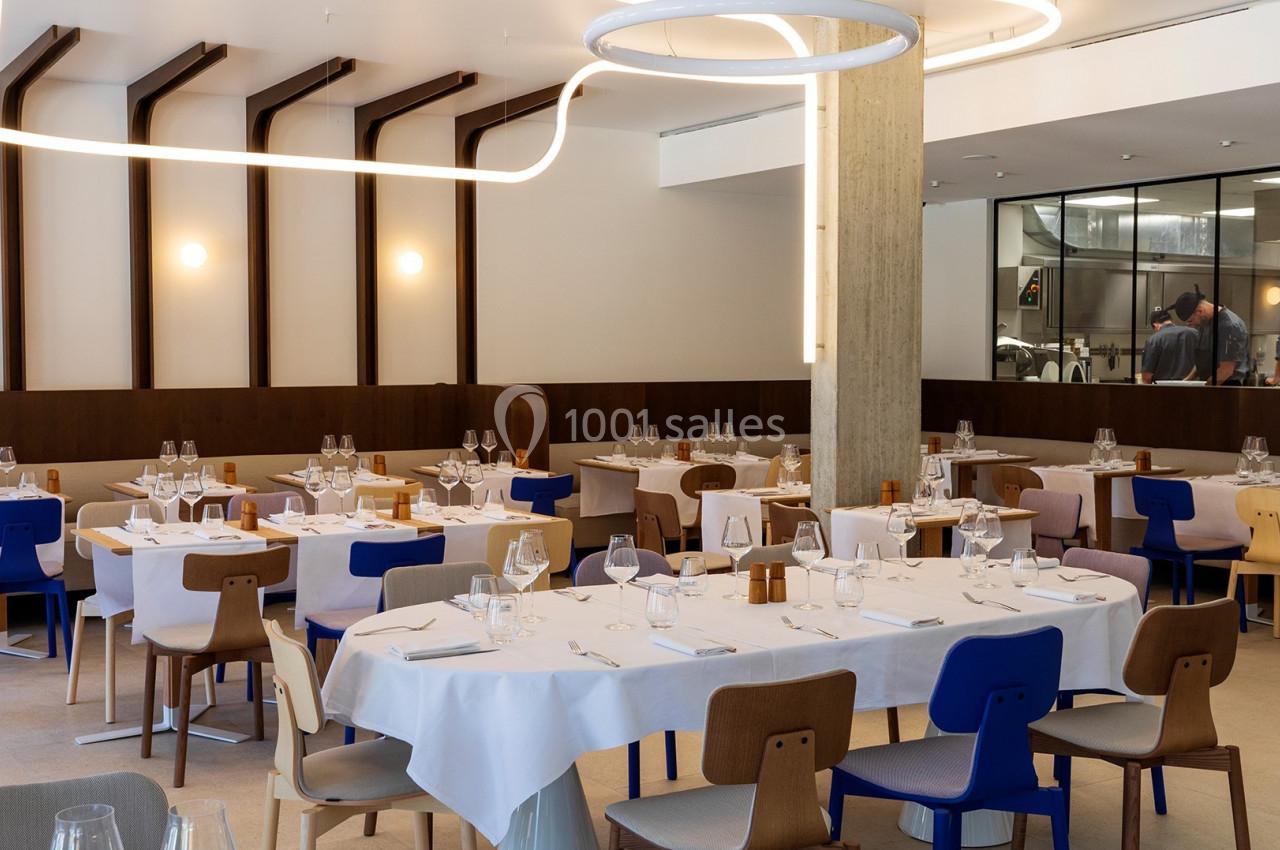 Salle de restaurant moderne avec tables dressées, chaises colorées et vue sur une cuisine ouverte à l'arrière.