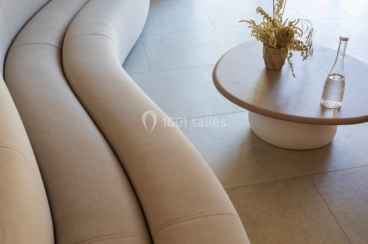 Canapé beige courbé près d'une table basse en bois clair avec un vase de fleurs séchées et une bouteille en verre.