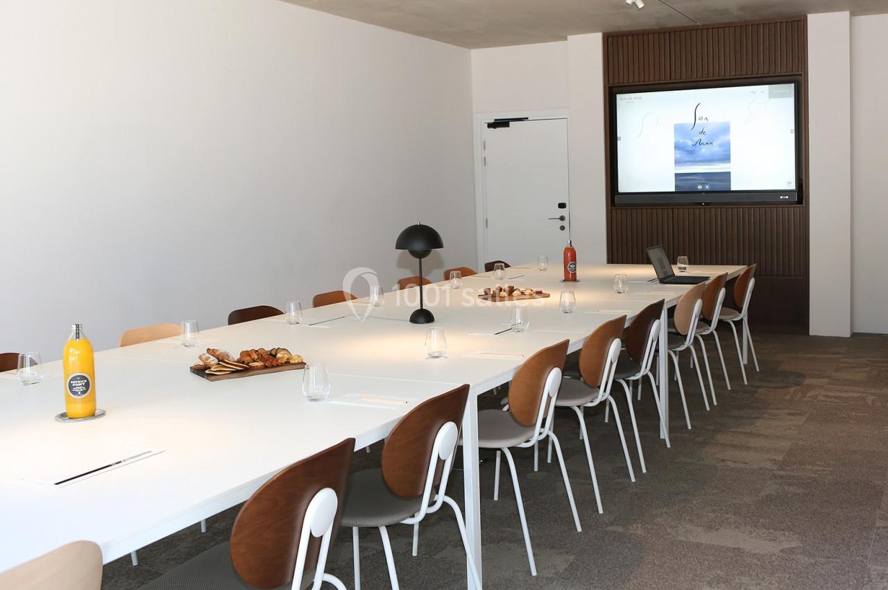 Salle de réunion moderne avec une grande table blanche, chaises en bois, écran mural et collations disposées.