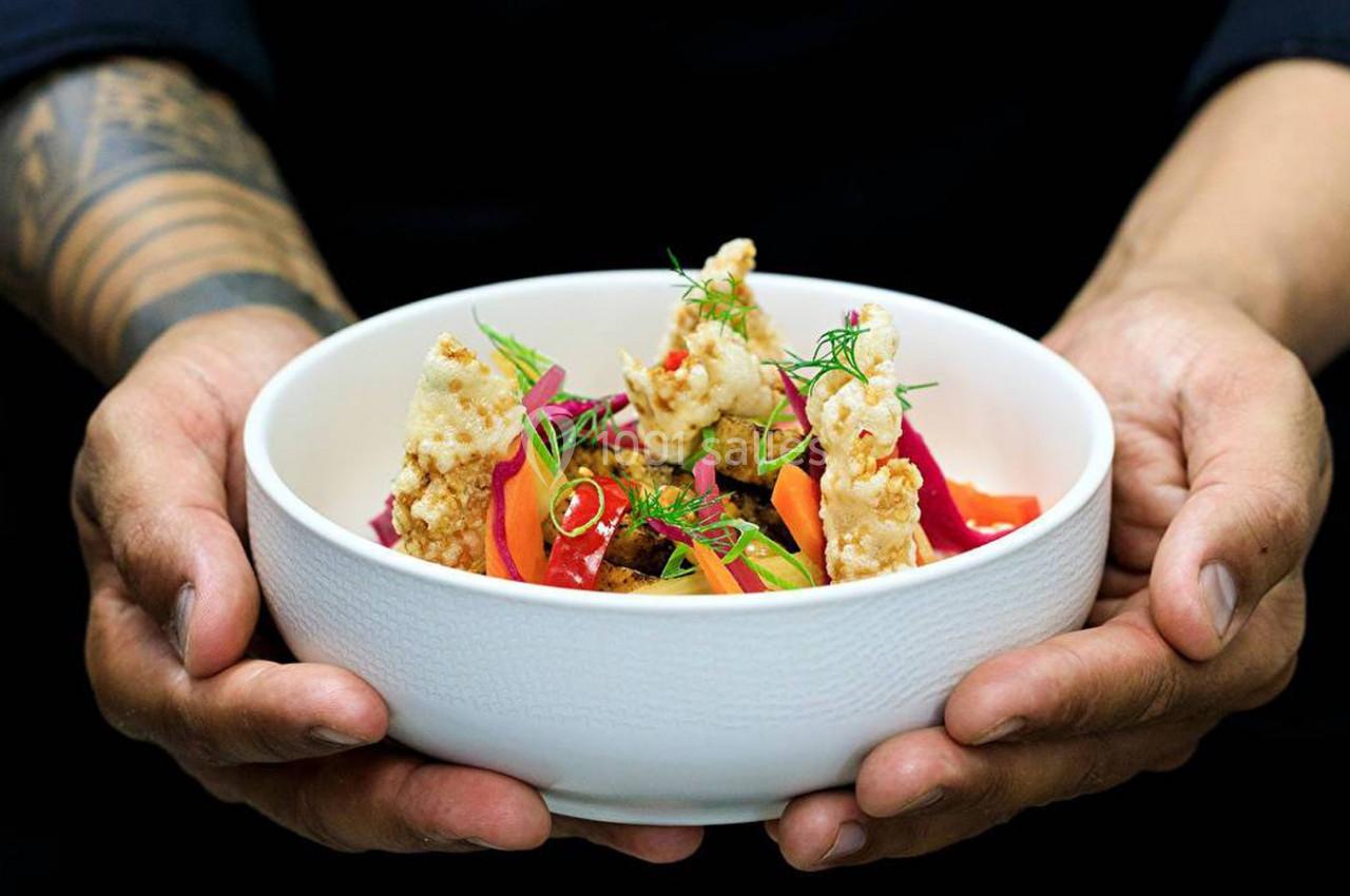 Bol blanc tenu par des mains, contenant un plat coloré avec légumes, herbes fraîches et morceaux croustillants.