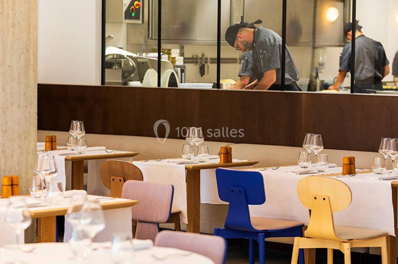 Salle de restaurant moderne avec tables dressées et vue sur une cuisine ouverte où travaillent des chefs.