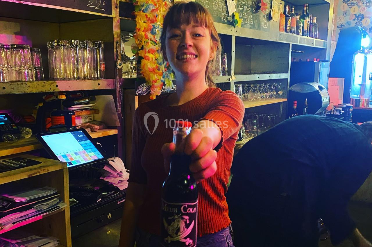 Jeune femme souriante tendant une bouteille de soda dans un bar éclairé, avec des étagères en arrière-plan.