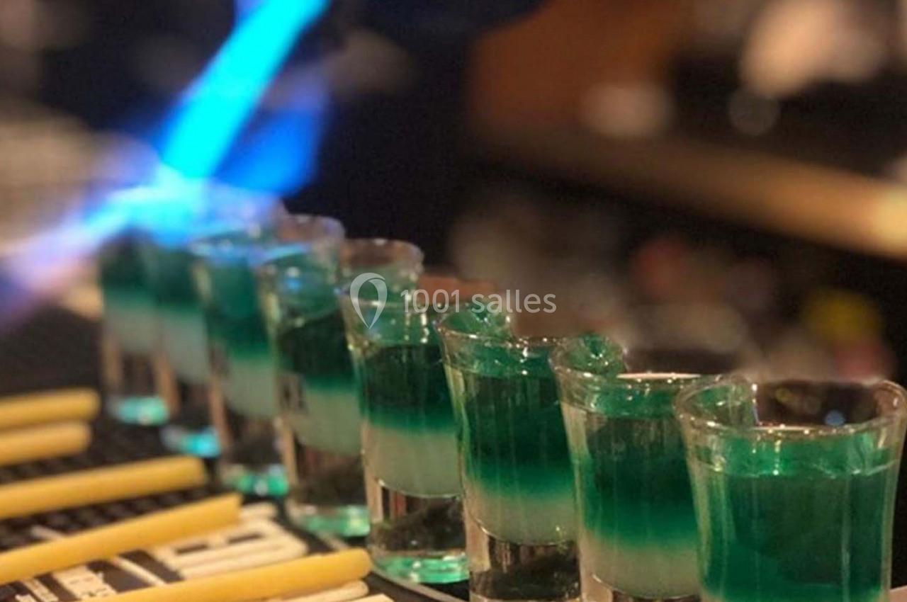 Verres à shooter alignés sur un comptoir, remplis d'un liquide vert et blanc, avec une flamme bleue en arrière-plan.