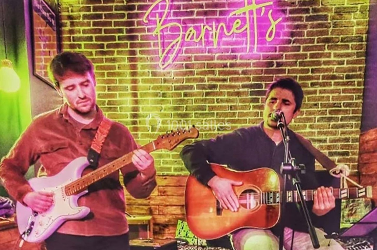 Deux musiciens jouent de la guitare devant un mur de briques avec une enseigne lumineuse en arrière-plan.