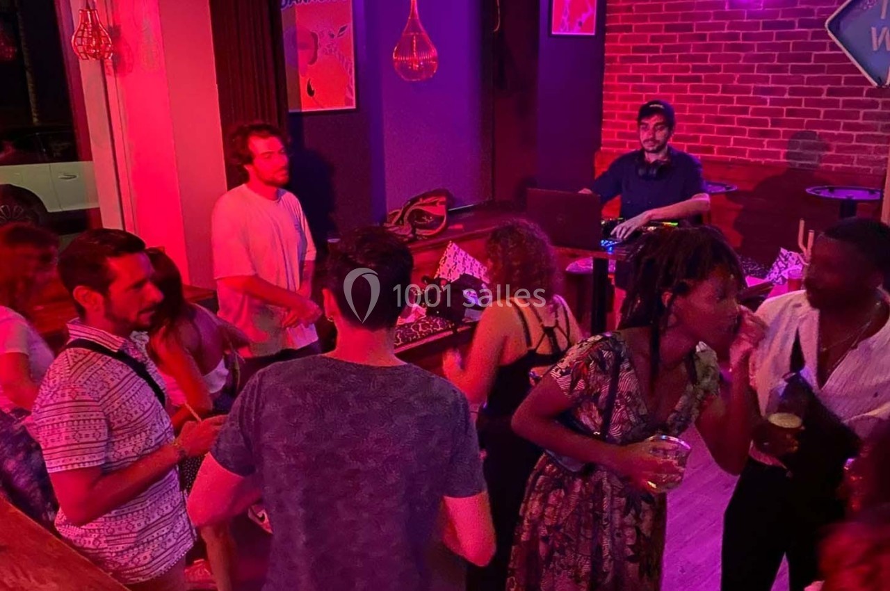 Des personnes dansent et discutent dans un bar à l'éclairage rouge, avec un DJ en arrière-plan.
