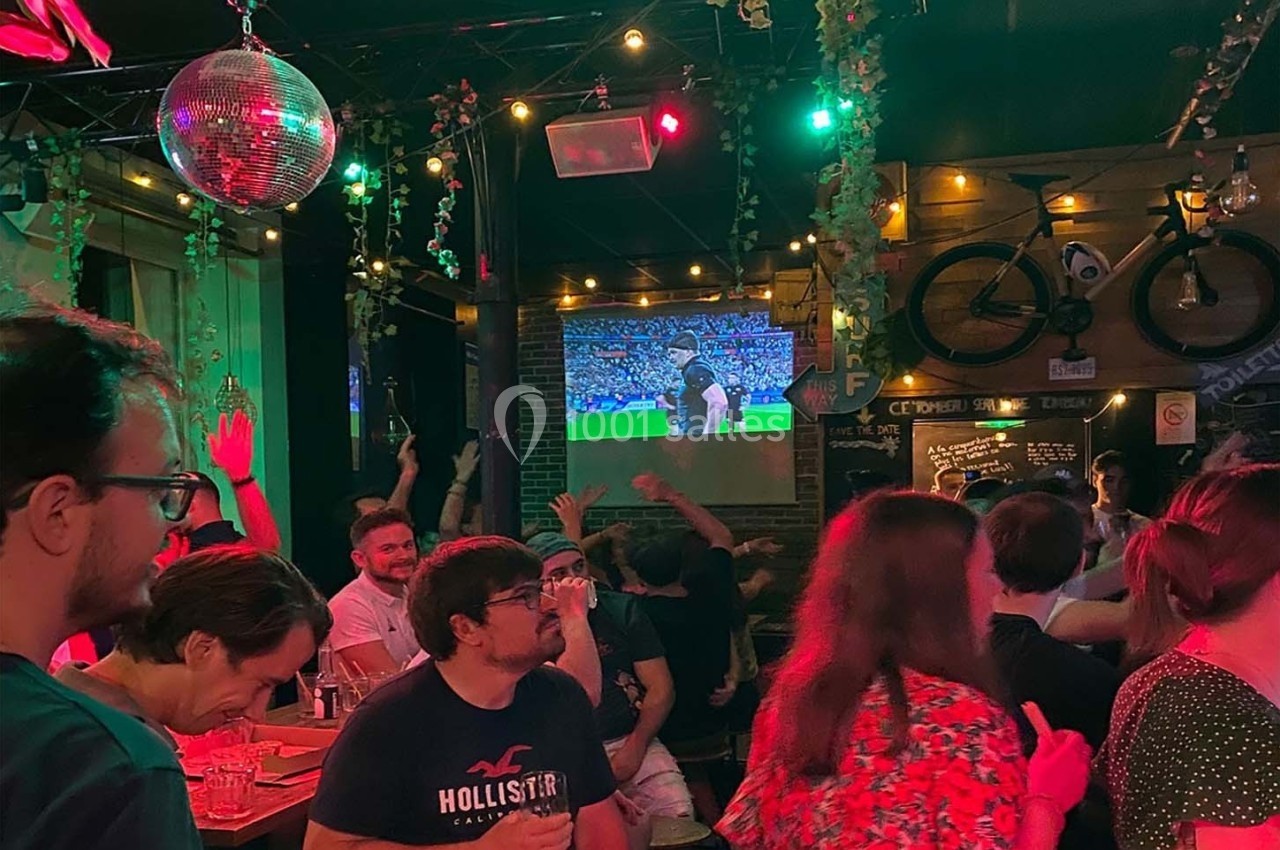 Des personnes regardent un match de football projeté dans un bar animé avec des lumières colorées et une ambiance festive.