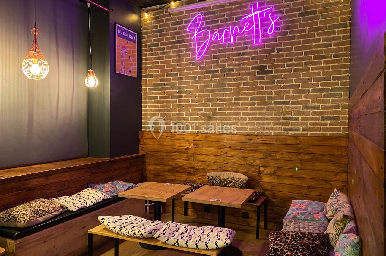 Coin salon cosy avec banquettes, coussins colorés, tables basses en bois et enseigne néon rose sur mur en briques.