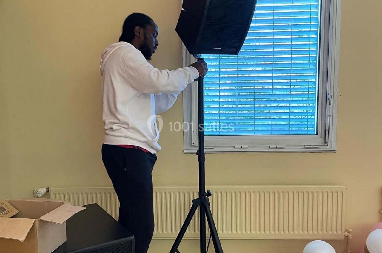 Un homme ajuste un haut-parleur sur un trépied dans une pièce avec des ballons et des cartons au sol.