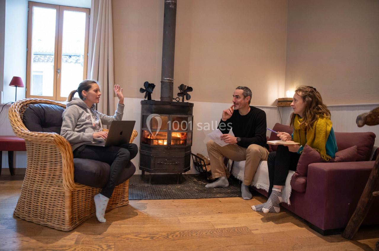 Trois personnes discutent dans un salon chaleureux près d'un poêle à bois, avec des fauteuils et une lumière naturelle.
