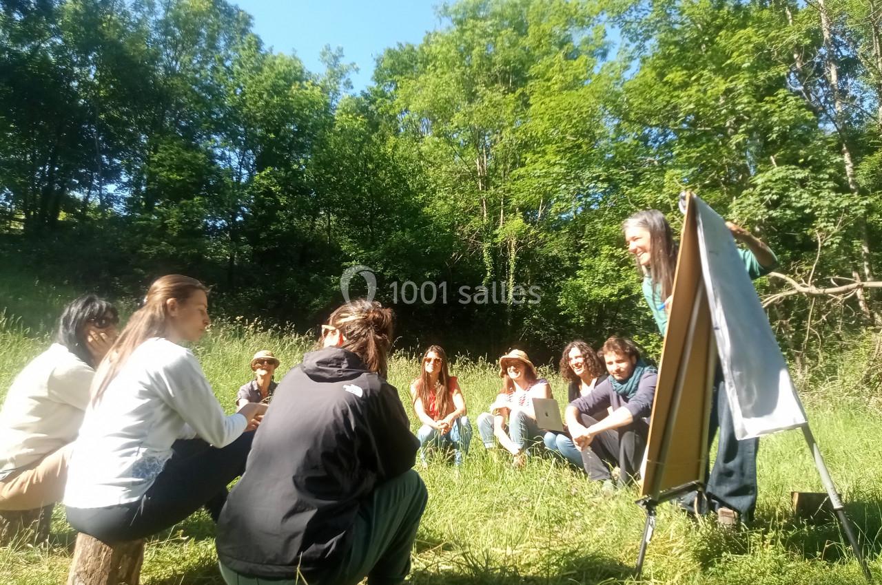 Un groupe de personnes assises en cercle dans un pré, écoutant une présentation devant un tableau chevalet.