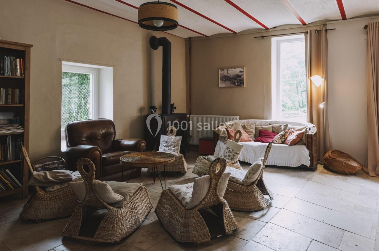 Salon chaleureux avec fauteuils en rotin, canapé, poêle à bois, bibliothèque et lumière naturelle.