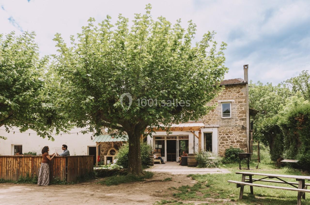 Maison en pierre entourée de verdure avec des arbres, une terrasse, des personnes discutant et une table de pique-nique.