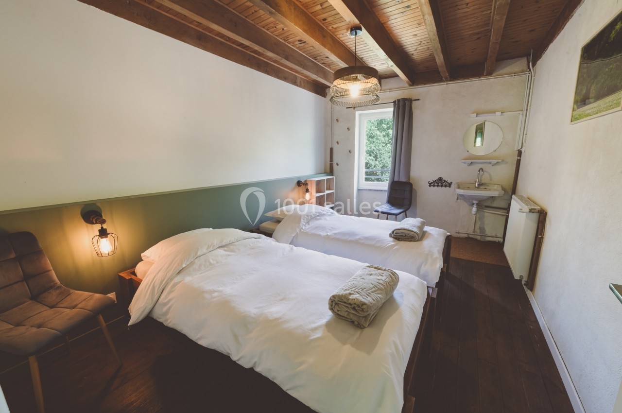 Chambre avec deux lits simples, plafond en bois, lavabo, fauteuil et fenêtre donnant sur un espace verdoyant.