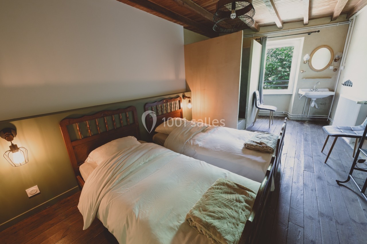 Chambre avec deux lits simples, parquet en bois, lavabo près d'une fenêtre donnant sur un espace verdoyant.