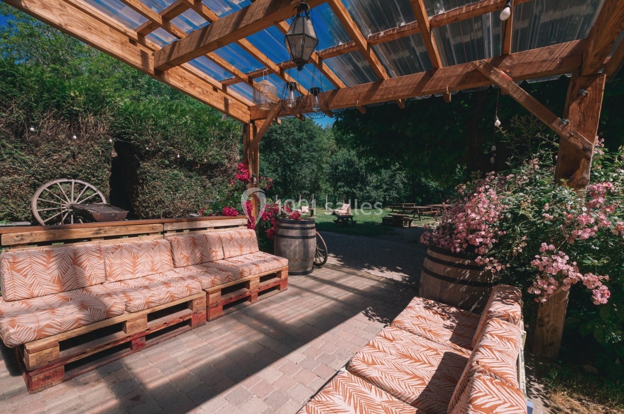 Terrasse en bois avec banquettes à motifs, fleurs roses, barriques et vue sur un jardin verdoyant en arrière-plan.