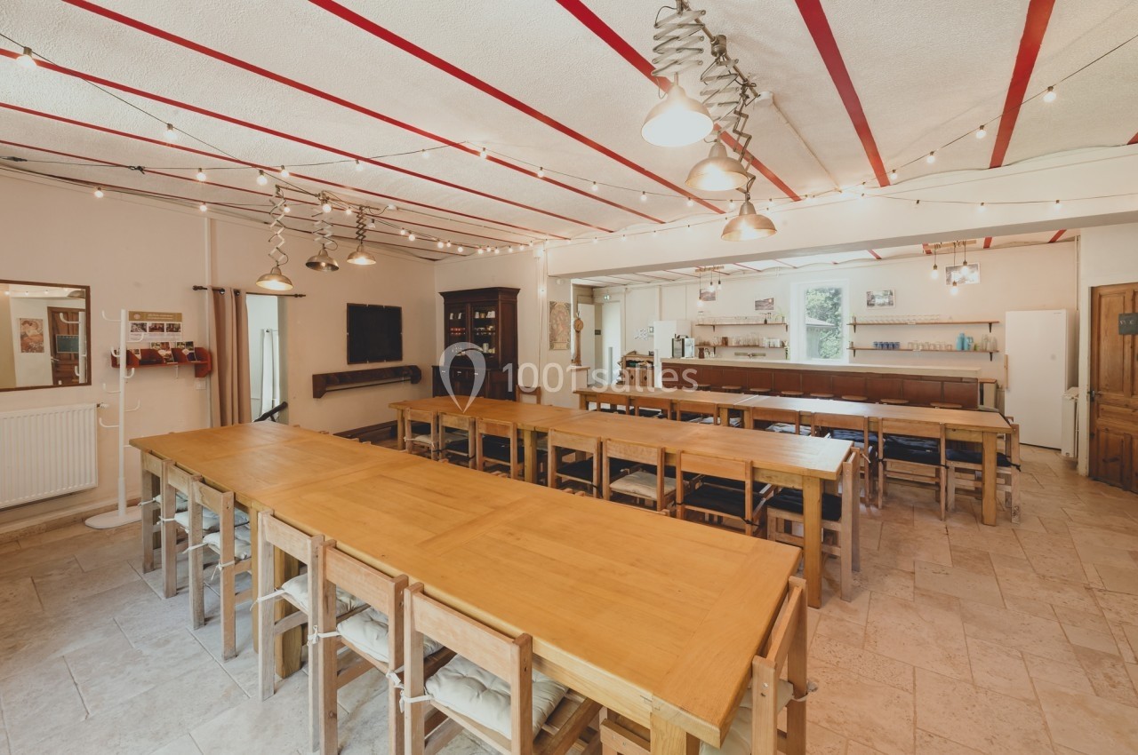Salle lumineuse avec de longues tables en bois, des chaises assorties et des guirlandes lumineuses au plafond.