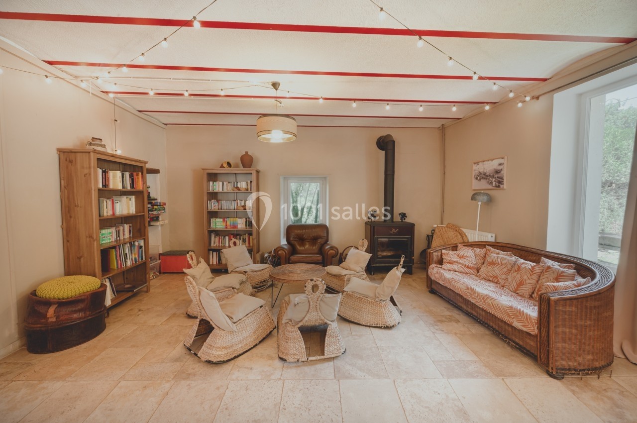 Salon chaleureux avec canapé en rotin, fauteuils en osier, bibliothèque, poêle à bois et guirlande lumineuse au plafond.