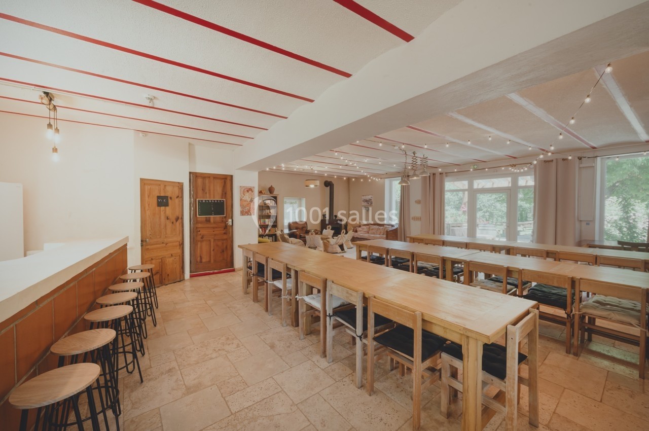 Salle lumineuse avec de longues tables en bois, des tabourets près d'un comptoir et des guirlandes lumineuses au plafond.