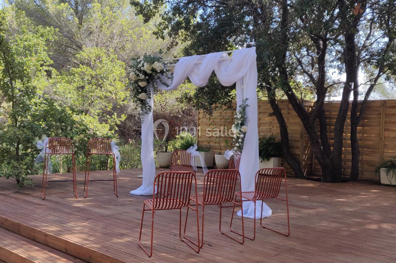 Arche de mariage décorée de voilages et de fleurs blanches sur une terrasse en bois entourée de végétation.