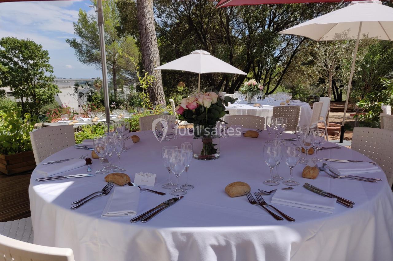 Table ronde dressée avec nappes blanches, verres, couverts et bouquet de roses, en terrasse ombragée avec vue sur jardin.