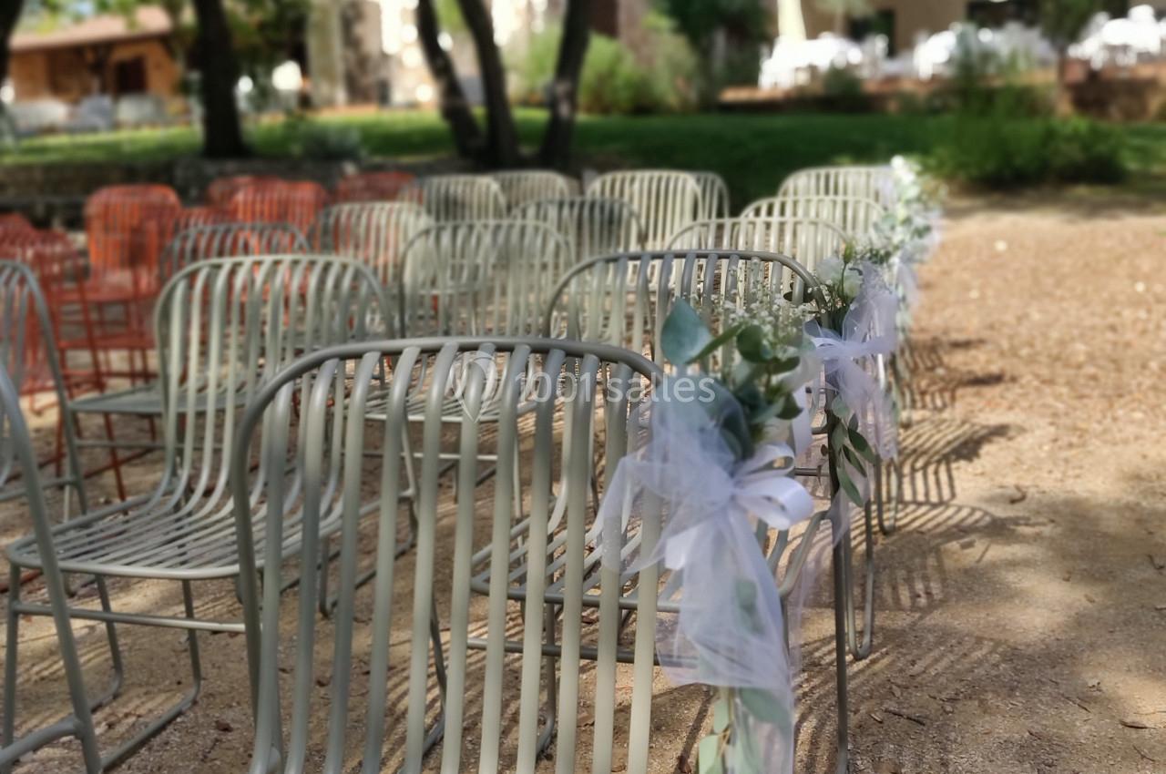 Chaises en métal alignées en extérieur, décorées de rubans blancs, dans un cadre arboré et ensoleillé.