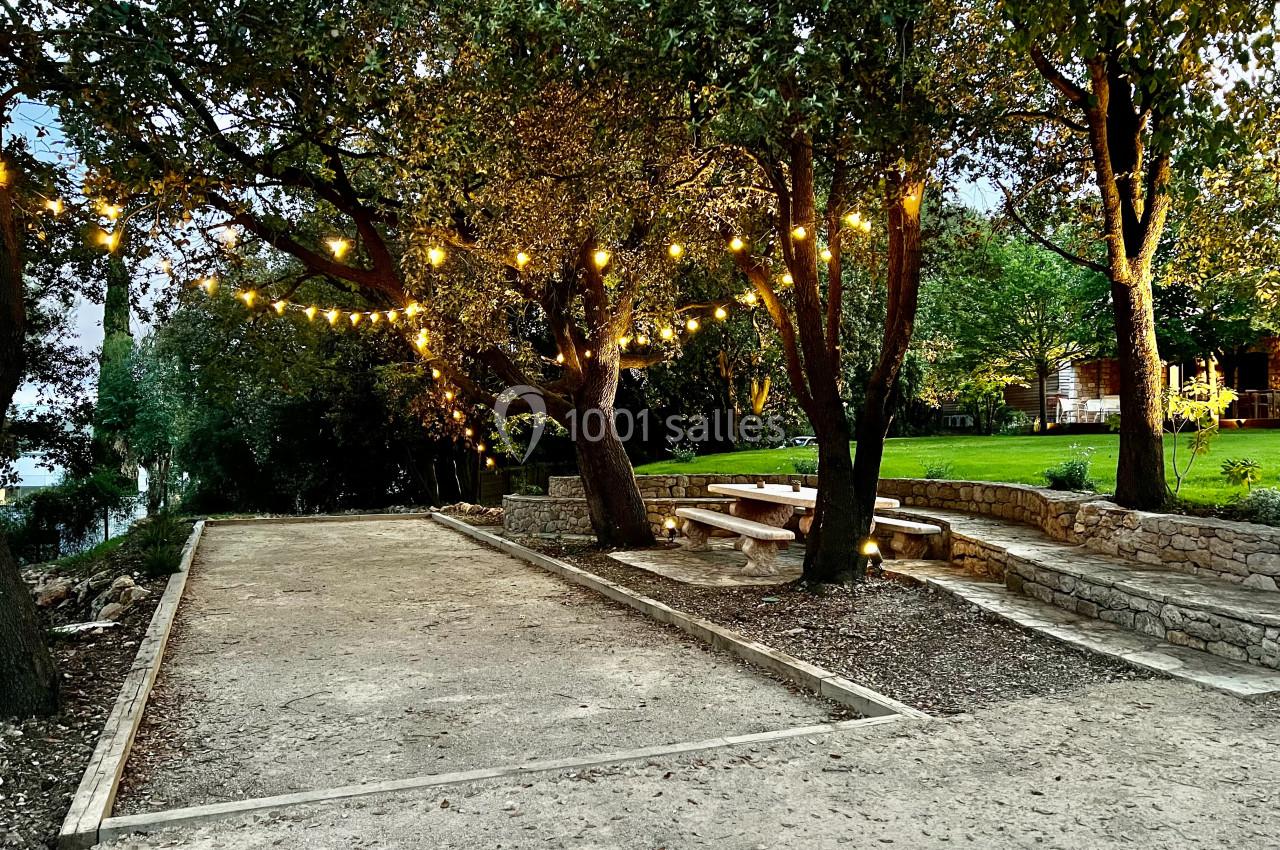 Terrain de pétanque entouré d'arbres, éclairé par des guirlandes lumineuses, avec une table de pique-nique en arrière-plan.
