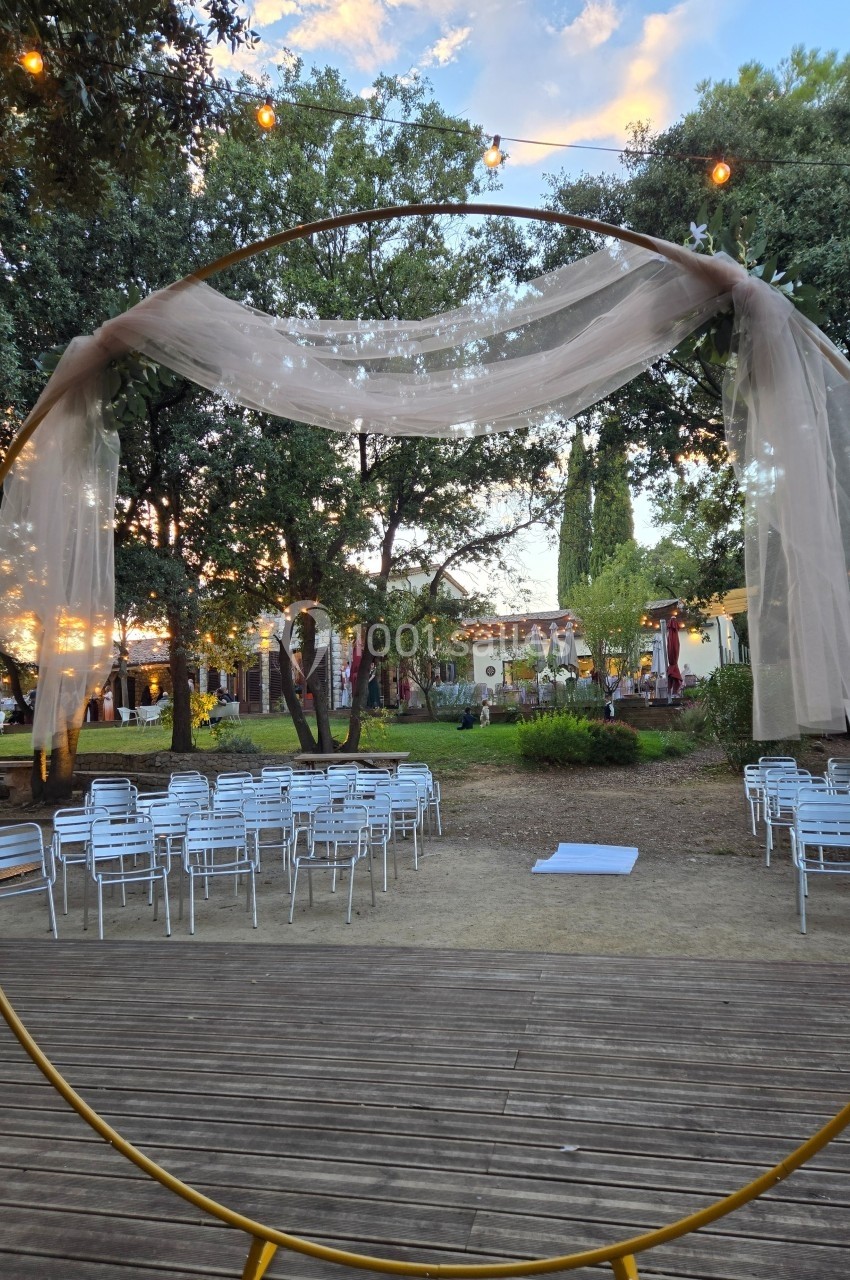Arche circulaire décorée de voilages, installée en extérieur devant des rangées de chaises et des arbres.