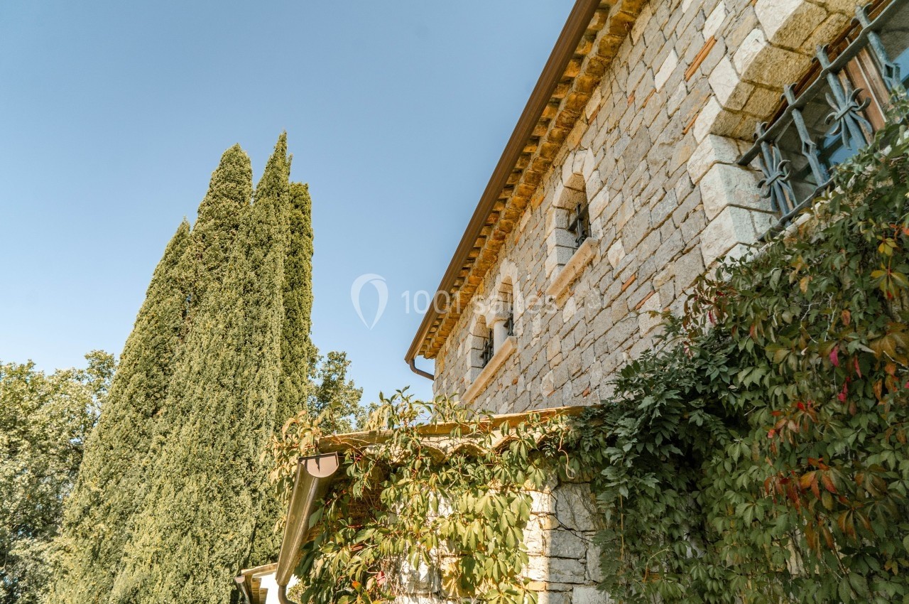 Façade en pierre d'une maison avec des volets fermés, entourée de végétation et d'un grand cyprès sous un ciel bleu.