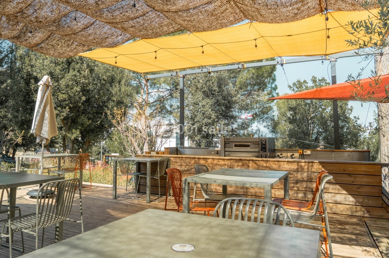 Terrasse extérieure aménagée avec des tables, des chaises, un barbecue et des voiles d'ombrage sous un ciel dégagé.