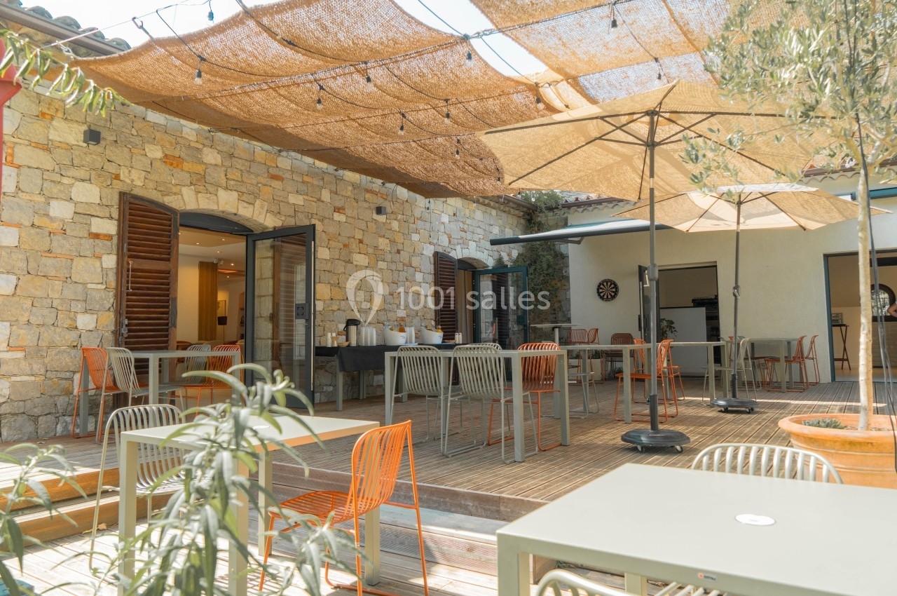 Terrasse ensoleillée avec tables, chaises colorées, parasols et mur en pierre, entourée de plantes.