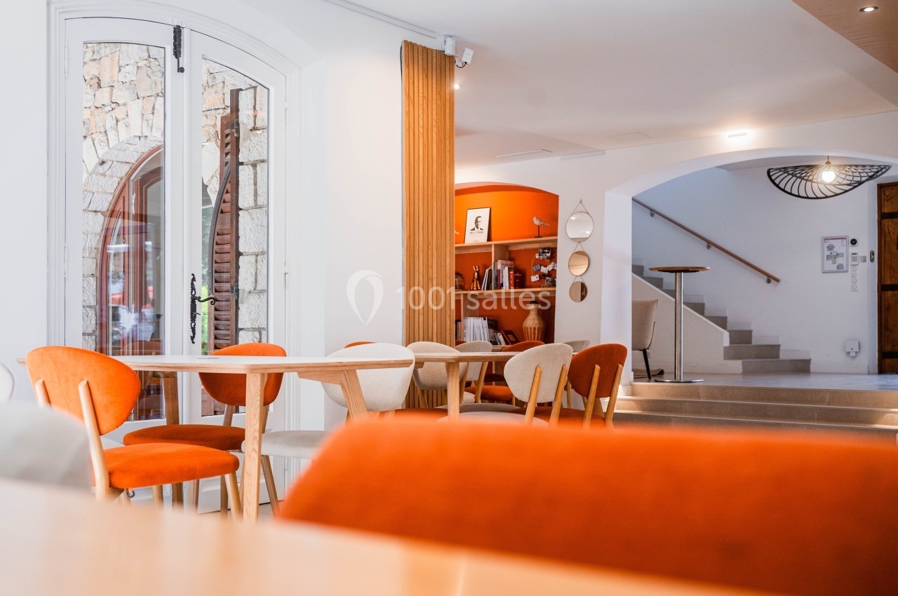Salle lumineuse avec des chaises orange et beige autour de tables en bois, escalier et étagères en arrière-plan.