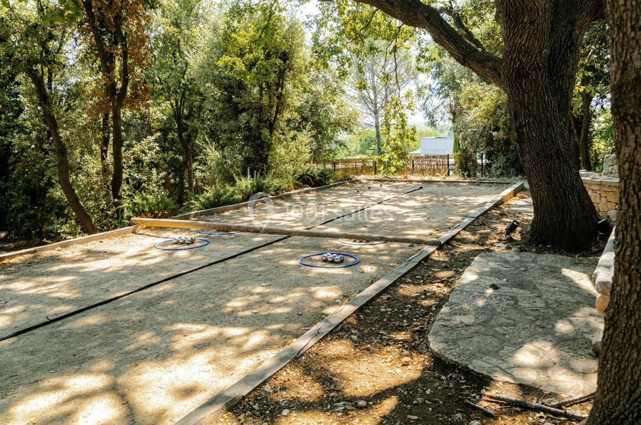 Terrain de pétanque ombragé entouré d'arbres, avec des cercles et des boules disposés au sol.