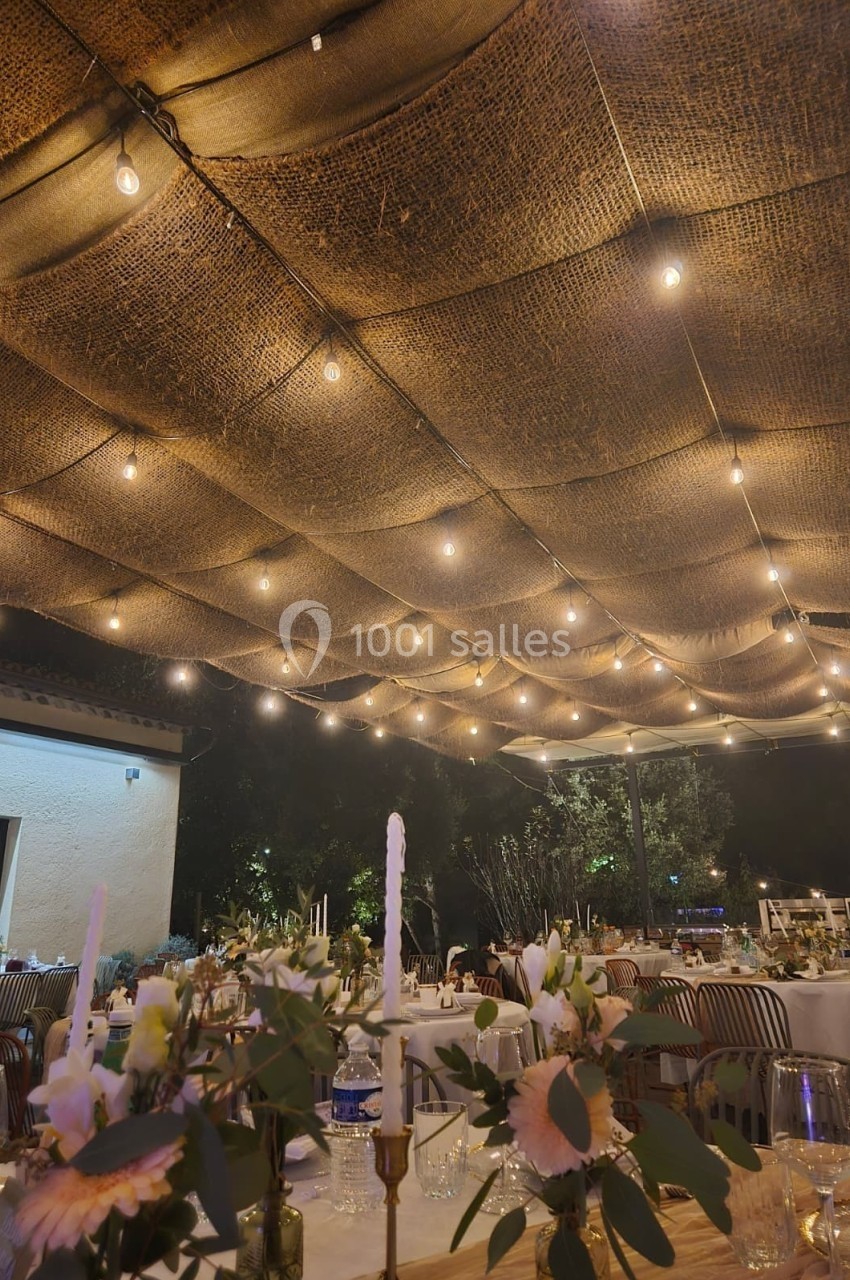 Décoration de table élégante sous un plafond de guirlandes lumineuses et de toile de jute en soirée extérieure.