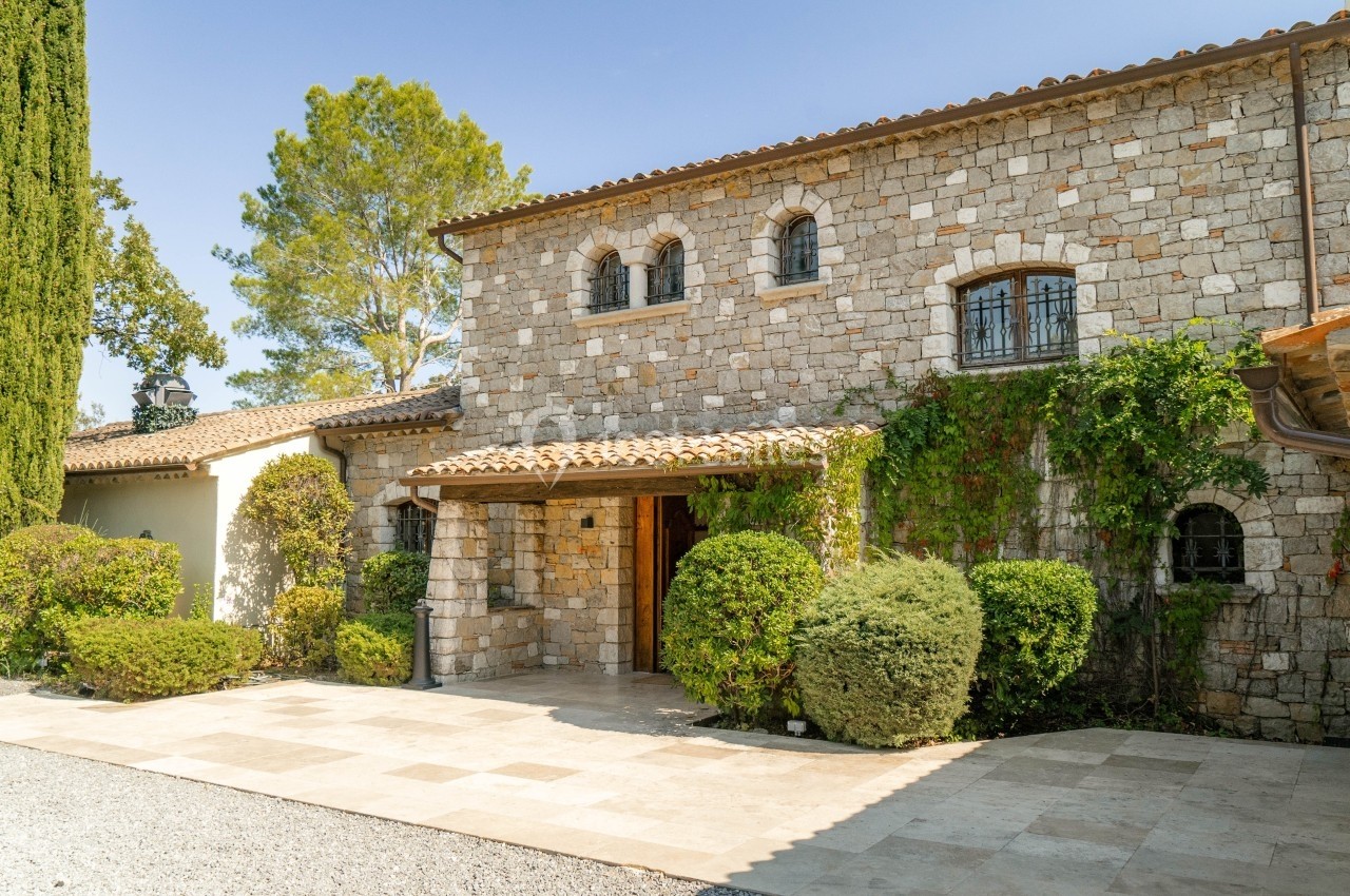 Façade en pierre d'une maison méditerranéenne entourée de végétation, avec une allée en pavés clairs.