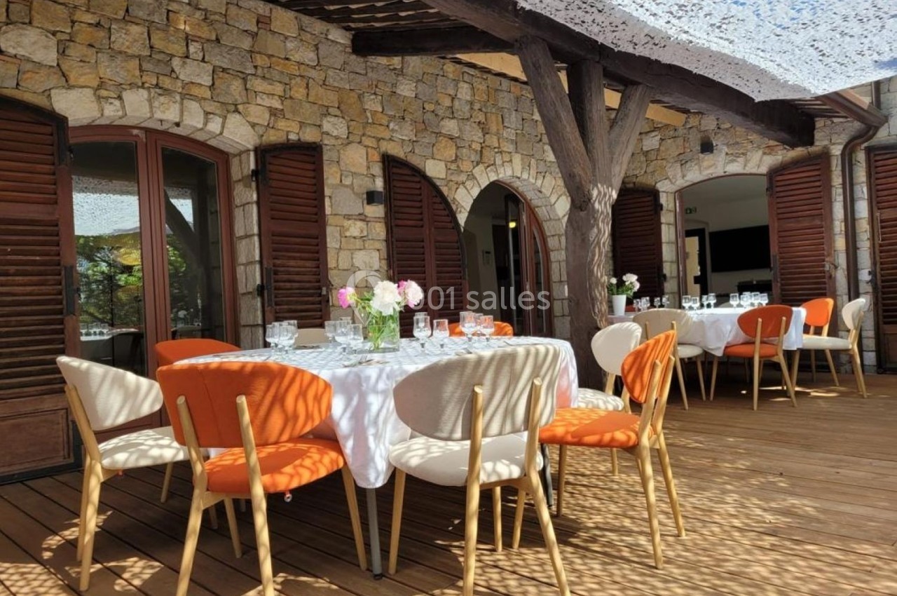 Terrasse en bois avec tables dressées, chaises colorées et ombrage partiel sous une pergola en pierre et bois.