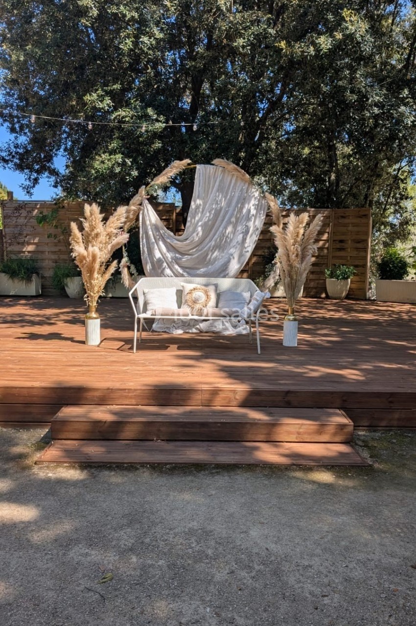 Banc décoré avec coussin et voilage beige, entouré de pampas, sur une scène en bois devant des arbres.