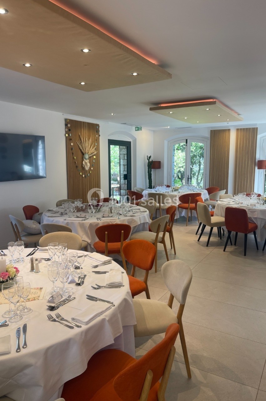 Salle de réception lumineuse avec tables rondes dressées, chaises colorées et grandes baies vitrées donnant sur un jardin.