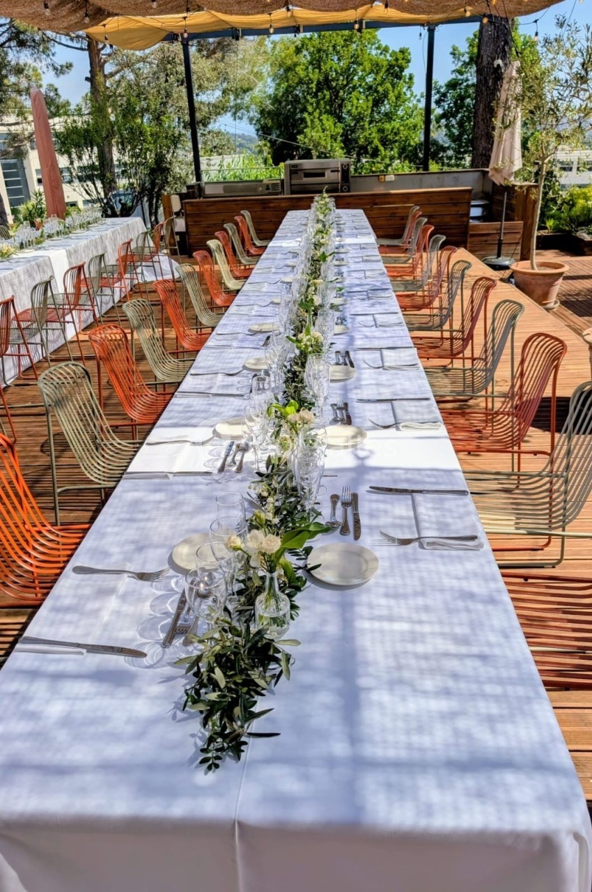 Table longue dressée en extérieur avec nappes blanches, vaisselle, verres et décoration florale centrale.