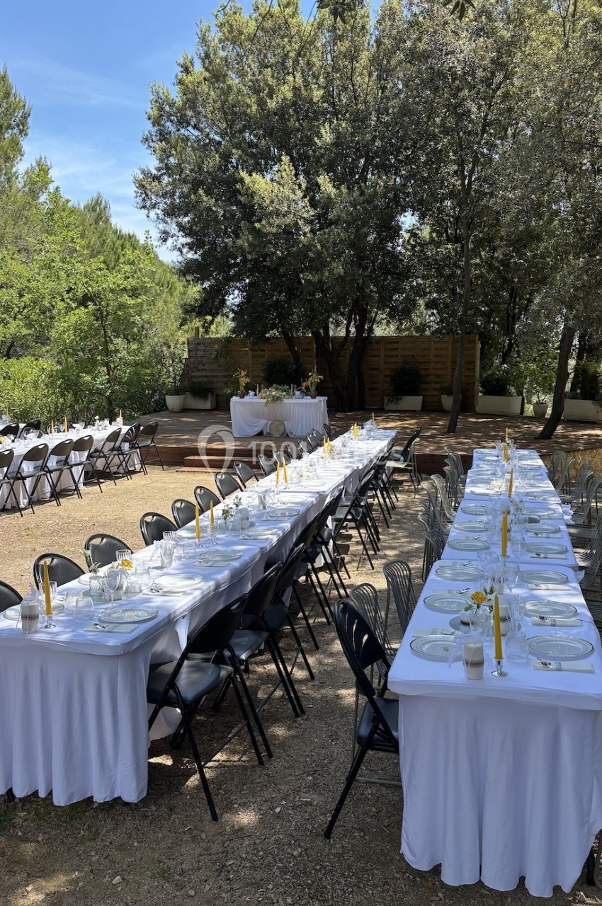 Tables dressées avec nappes blanches, assiettes et bougies jaunes, disposées en extérieur sous des arbres.