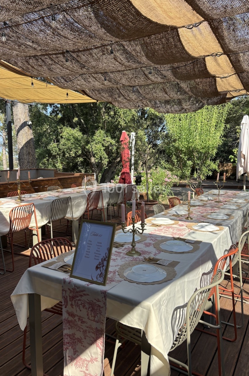 Tables dressées en extérieur sous une pergola ombragée, avec nappes décoratives, vaisselle et chandeliers.
