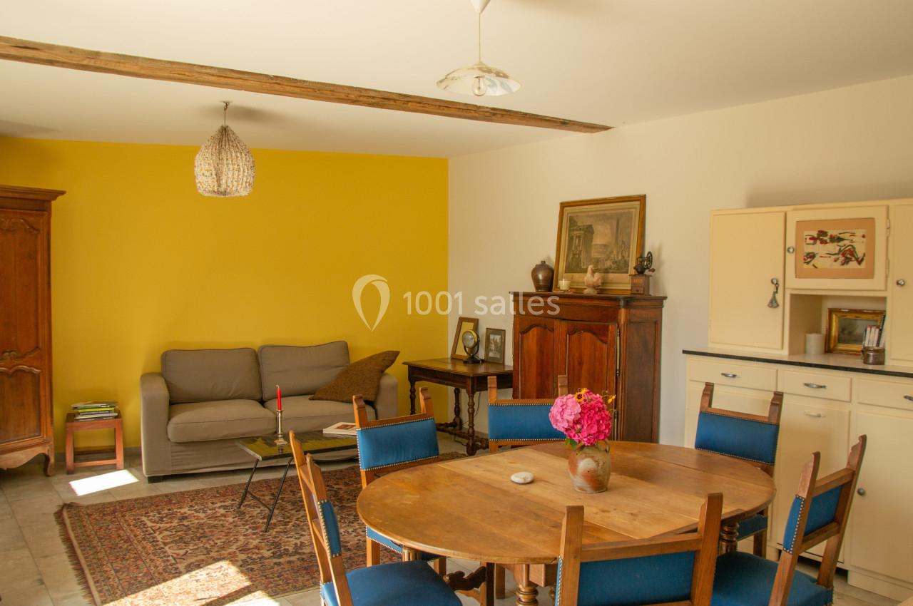 Salle à manger lumineuse avec table en bois, chaises bleues, canapé gris, mur jaune et meubles anciens.