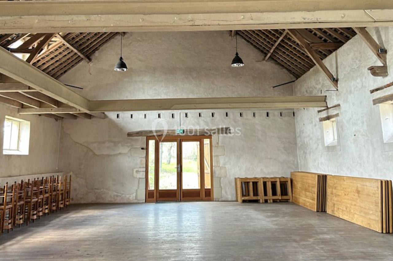 Salle vide avec murs en pierre blanche, poutres apparentes, parquet en bois et chaises empilées sur le côté.