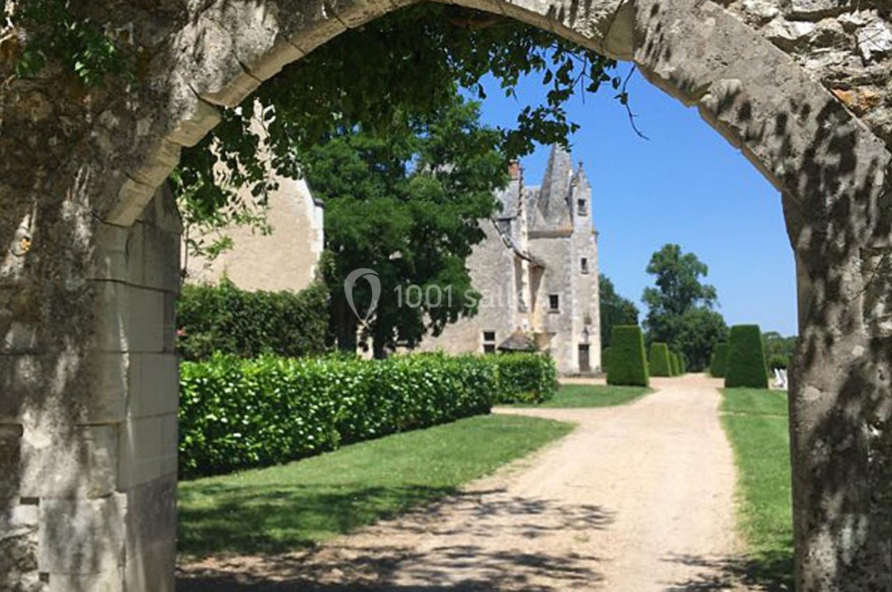 Vue d'une allée bordée de verdure menant à un bâtiment en pierre sous une arche en pierre ancienne.
