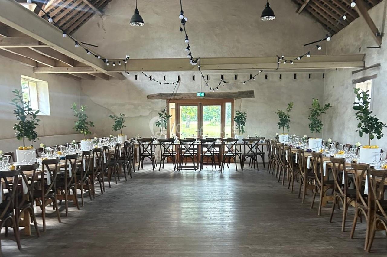 Salle de réception rustique avec tables en bois dressées, guirlandes lumineuses suspendues et grandes fenêtres donnant sur l…