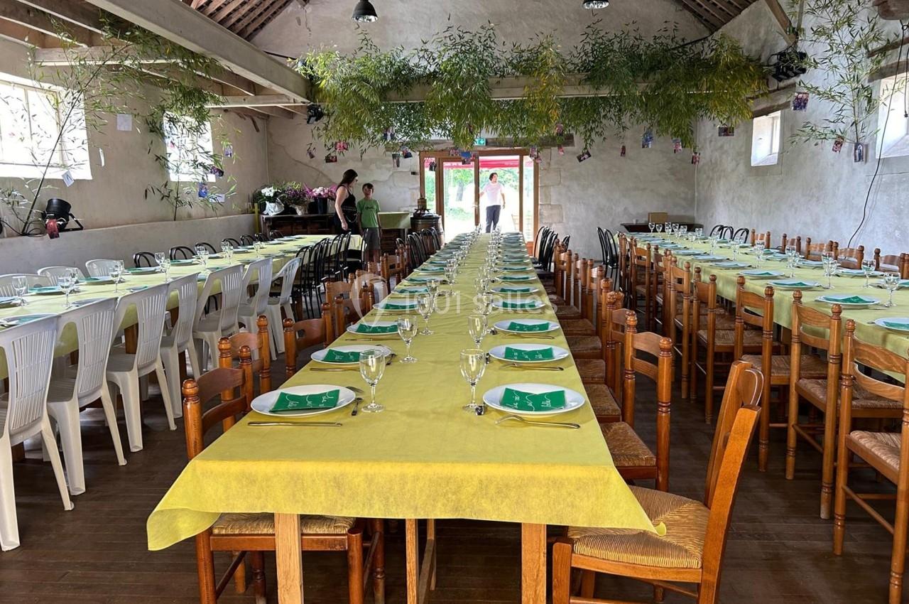 Salle de réception avec deux longues tables dressées, nappes jaunes, assiettes, verres et décorations végétales suspendues.